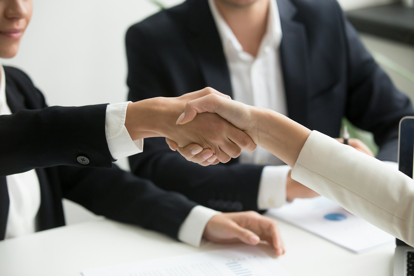 Two business professionals shake hands over a desk in an office.